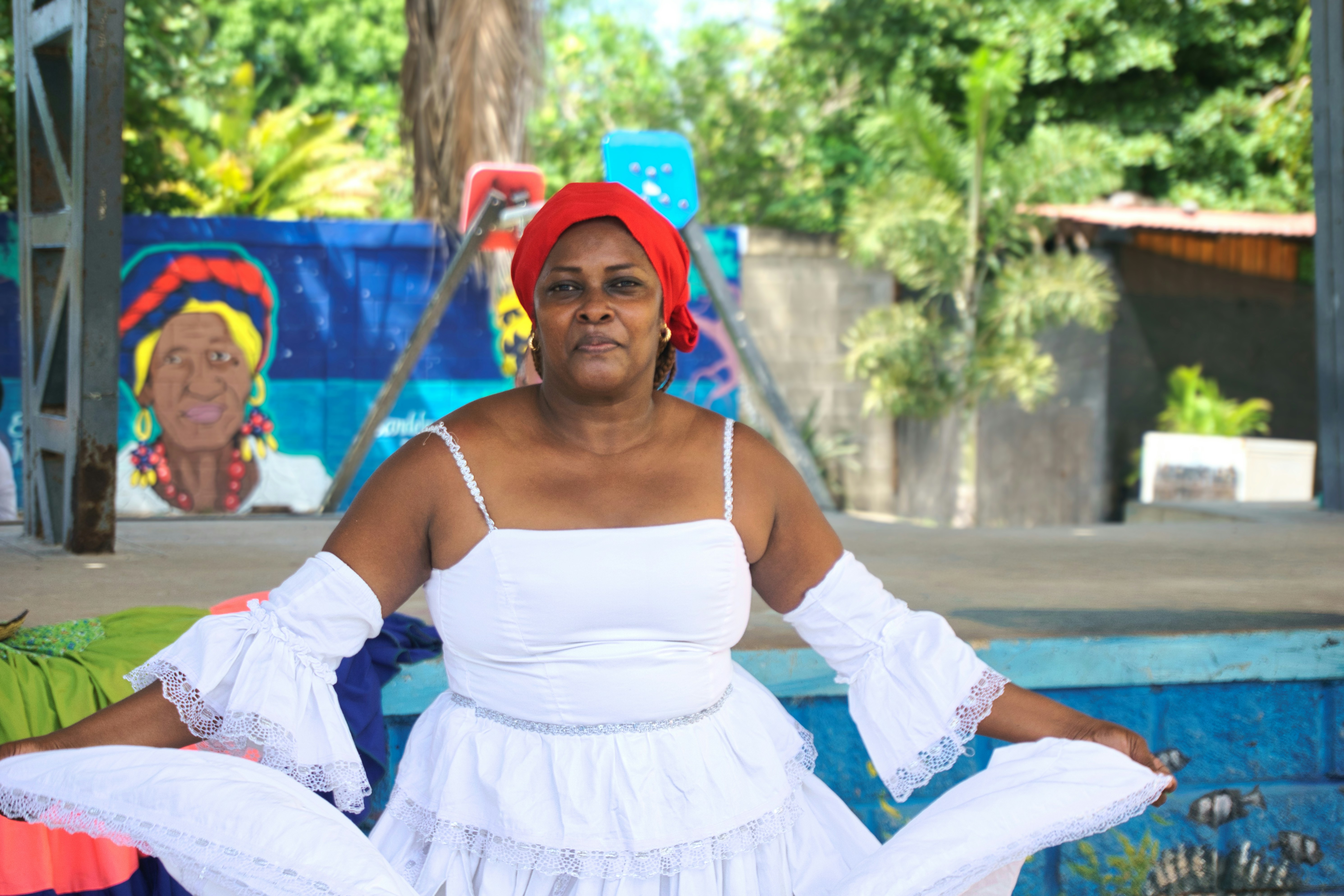 A dignified portrait of a woman wearing a vibrant red headwrap, positioned in front of a colorful mural that pays tribute to Afro-descendant identity in Santa Ana, Barú. The natural light highlights the strength of her expression.